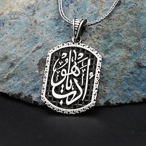 May include: A silver pendant necklace with an ornate rectangular pendant. The pendant features Arabic calligraphy in a white, stylized font set against a black grid background. The necklace chain is a fine, textured silver.