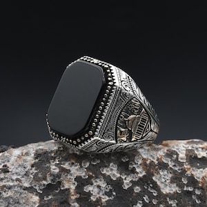 May include: A silver ring with a rectangular black onyx stone. The ring features intricate engraved patterns and gold accents on the sides. The ring is displayed on a textured gray rock against a dark background.