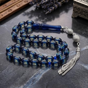 May include: A string of prayer beads featuring translucent blue oval beads with a yellow core. The strand includes a long blue focal bead, two silver-toned textured spherical beads, and a silver-toned metal tassel at the end of the chain.