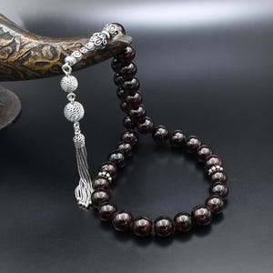 Peut inclure: Un chapelet de perles de pierres précieuses polies, probablement grenat, de couleur rouge foncé, avec des éléments décoratifs argentés. Les perles sont enfilées ensemble, formant une boucle. Les composants argentés comprennent des entretoises ornées et un pompon.