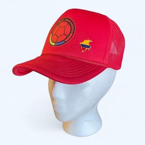 Puede incluir: Gorra de camionero roja con visera curva y parte trasera de malla. La gorra presenta un diseño de balón de fútbol con los colores de la bandera colombiana y un pin del mapa colombiano. El texto en la gorra dice "COLOMBIA PARA EL MUNDO HINCHA".