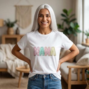 May include: A white t-shirt with the word "MAMA" in pastel colors. The letters are in a patchwork style with pink, green, and yellow patterns. The shirt is worn by a person with silver hair and blue jeans.