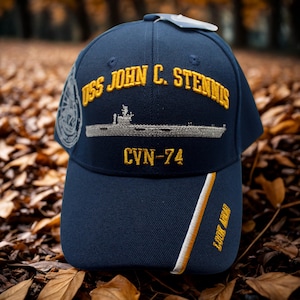 Könnte beinhalten: Marineblaue Baseballkappe mit goldgesticktem Text "USS JOHN C. STENNIS" und "CVN-74". Mit einer silbernen Illustration eines Flugzeugträgers. Die Kappe hat einen weißen und goldenen Streifen an der Seite und den Text "LOOK AHEAD".