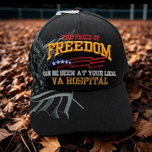 May include: Black baseball cap with embroidered text "THE PRICE OF FREEDOM" in yellow and red, with an American flag design. The text "CAN BE SEEN AT YOUR LOCAL VA HOSPITAL" is also embroidered on the cap. The cap has a curved brim.