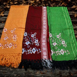 May include: Three colorful scarves with floral embroidery and fringe. The scarves are in shades of orange, burgundy, and green. The orange scarf has orange fringe, the burgundy scarf has black fringe, and the green scarf has black fringe.