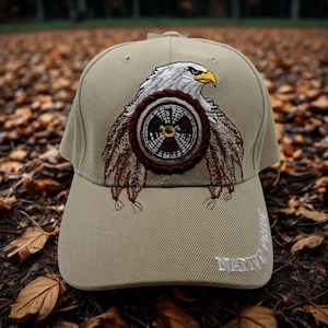 Puede incluir: Una gorra de béisbol de color tostado con un diseño bordado de un águila blanca con pico amarillo sobre un emblema circular con patrones marrones y blancos. El texto NATIVE PRIDE está bordado en blanco en el lado inferior derecho de la visera.