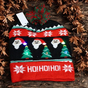 May include: A Christmas beanie in red, white, and black, featuring Santa Claus faces and Christmas trees with LED lights. The brim displays the text "HO! HO! HO!" and snowflakes. A red and green pom-pom tops the hat. The hat is on a wooden surface.