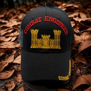 May include: Black baseball cap with "COMBAT ENGINEER" embroidered in red and gold. The cap features a gold castle emblem and additional text on the side. The hat is set against a backdrop of fallen leaves.