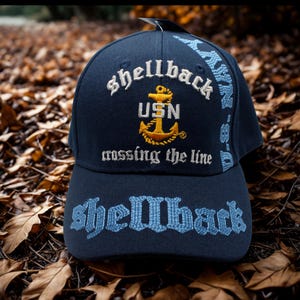 May include: Navy blue baseball cap with embroidered text and a gold anchor emblem. The cap features the words "Shellback", "USN", and "crossing the line" in white and gold thread. The brim also has the word "Shellback" in light blue.