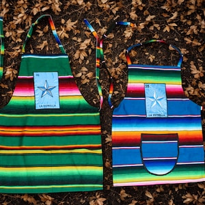 May include: Two colourful aprons with a striped design in green and blue. Each apron features a light blue patch with a star and the text "LA ESTRELLA". The aprons are made of a woven fabric and have long ties.