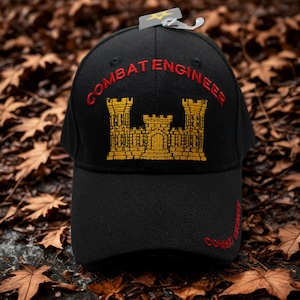 May include: Black baseball cap with "COMBAT ENGINEER" embroidered in red and a gold castle emblem. The cap has a curved brim and a tag attached. The text "COMBAT ENGINEER" is also embroidered on the side.