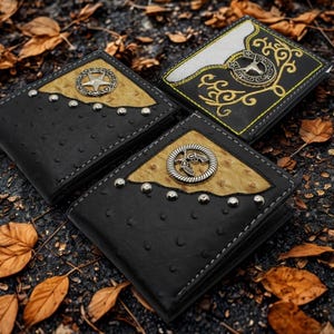 May include: Three black leather wallets with decorative silver accents and a light brown panel. One wallet features a star, another a horseshoe, and the third a revolver design. The wallets are arranged on a surface with fallen leaves.