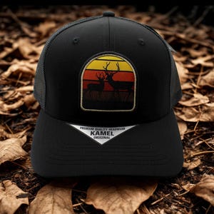 Pode incluir: Boné de caminhoneiro preto com um patch com duas silhuetas de veados contra um fundo de pôr do sol amarelo, laranja e vermelho. O boné tem uma aba curva e uma parte traseira de malha. O texto "Premium Quality Headwear KAMEL Original" é visível.