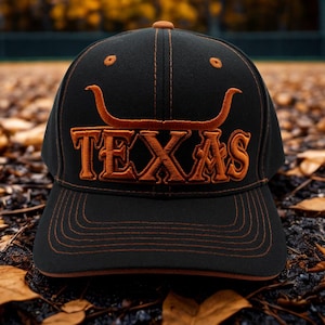 May include: Black baseball cap featuring the word "TEXAS" in orange, and an orange longhorn symbol. The cap has a curved peak and is set against a backdrop of autumn leaves.