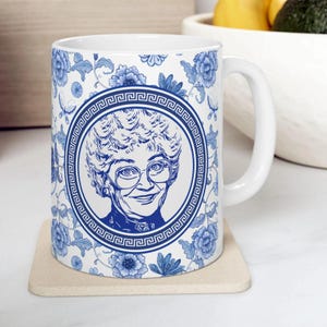 May include: A white ceramic mug featuring a blue floral pattern and a central circular medallion containing a line-art portrait of an older person with short curly hair and glasses, framed by a Greek key border design.