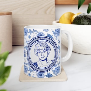 May include: White ceramic mug with a blue and white floral pattern. The mug features a portrait of a woman in a circle with a blue and white Greek key border.