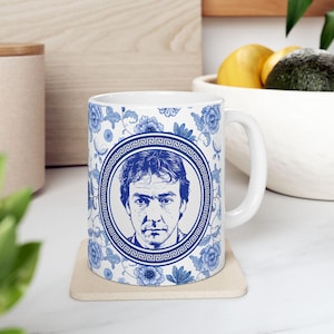 May include: A white ceramic mug with a blue and white design. The mug features a portrait of a man in a circle with a Greek key border. The design also includes floral patterns.