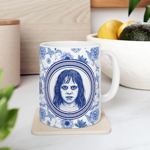 May include: White ceramic mug with a blue floral pattern and a central portrait of a person with a stern expression. The portrait is framed by a Greek key border. The mug has a white handle.