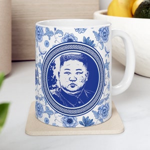 May include: White ceramic mug with a blue and white design. The design features a portrait of a man in a circle with a blue and white patterned border. The background is a white floral pattern.