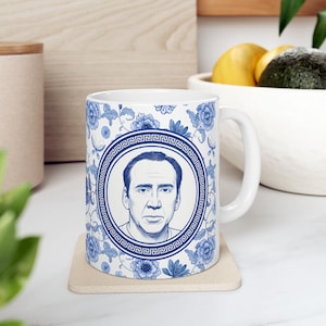 May include: White ceramic mug featuring a portrait of Nicolas Cage in a blue and white floral design. The mug has a white handle and is placed on a beige coaster. The design includes a Greek key border.
