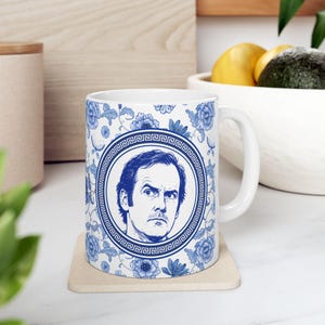 May include: A white ceramic mug featuring a blue and white floral pattern with a central circular medallion containing a detailed blue illustration of a man's face. The medallion is framed by a geometric Greek key border design.