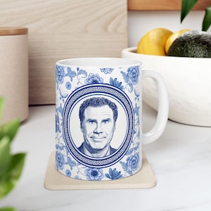 May include: White ceramic mug featuring a portrait of a man in a blue and white floral design. The portrait is framed by a Greek key border. The mug is on a beige coaster.