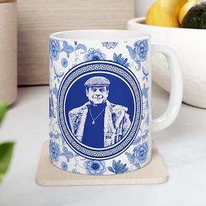 May include: A white ceramic mug with a blue and white floral pattern. The mug features a portrait of a man wearing a blue cap and a blue jacket. The portrait is surrounded by a blue and white Greek key border.