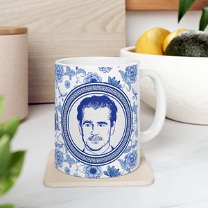 May include: A white ceramic mug with a blue and white floral pattern. The mug features a portrait of a man in a circle with a Greek key border. The mug is on a light brown coaster.