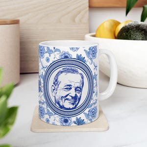 May include: White ceramic mug featuring a blue and white floral pattern and a portrait of a smiling person. The portrait is framed by a Greek key border. The mug sits on a beige coaster.