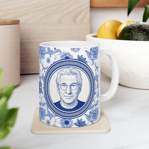 May include: White ceramic mug with a blue and white floral pattern and a portrait of a man in a circular frame. The mug has a white handle and sits on a beige coaster.