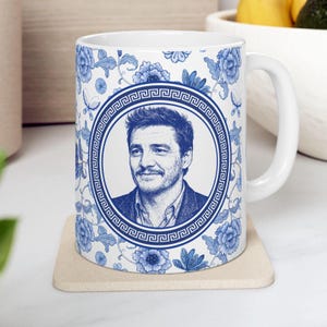 May include: White ceramic mug featuring a portrait in a blue and white floral design. The portrait is framed by a Greek key border. The mug has a white handle and is sitting on a beige coaster.
