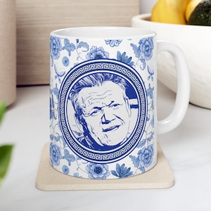 May include: White ceramic mug with a blue and white floral pattern. A blue circle with a Greek key border surrounds a black and white portrait of a man.