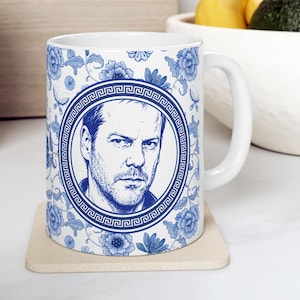 May include: A white ceramic mug featuring a blue and white floral pattern with butterflies. Centered on the mug is a circular frame with a Greek key border containing a detailed blue line drawing of a man with short hair and stubble.