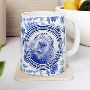 May include: White ceramic mug with a blue and white design. The design features a stylized beaver head in a circle with a Greek key border. The background is a floral pattern with blue flowers and leaves.