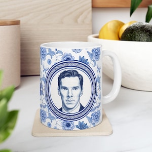 May include: White ceramic mug featuring a portrait in a blue and white floral design. The portrait is framed by a Greek key border. The mug has a white handle and is placed on a beige coaster.