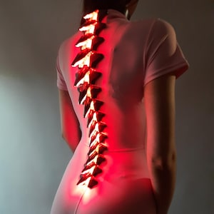 May include: A pink jumpsuit with a glowing red spine-like structure down the back. The spine is made of angular, light-emitting pieces. The person is facing away from the camera, with the jumpsuit's design emphasizing the spine's illumination.