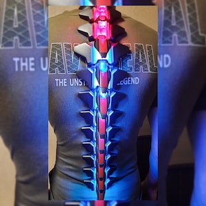 May include: A person wearing a dark grey t-shirt with the text "The Unstoppable Legend" printed on it. The person is wearing a black and blue light-up spine accessory with red lights.