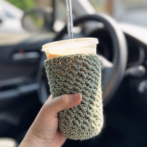 May include: A hand-crocheted, olive green coffee cup cozy. The cozy is wrapped around a clear plastic cup filled with a beverage. The background shows a car interior, including the steering wheel and dashboard.
