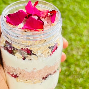 May include: A glass jar filled with layers of pink Himalayan salt, oatmeal, and rose petals. The jar is held in a hand against a green grassy background.