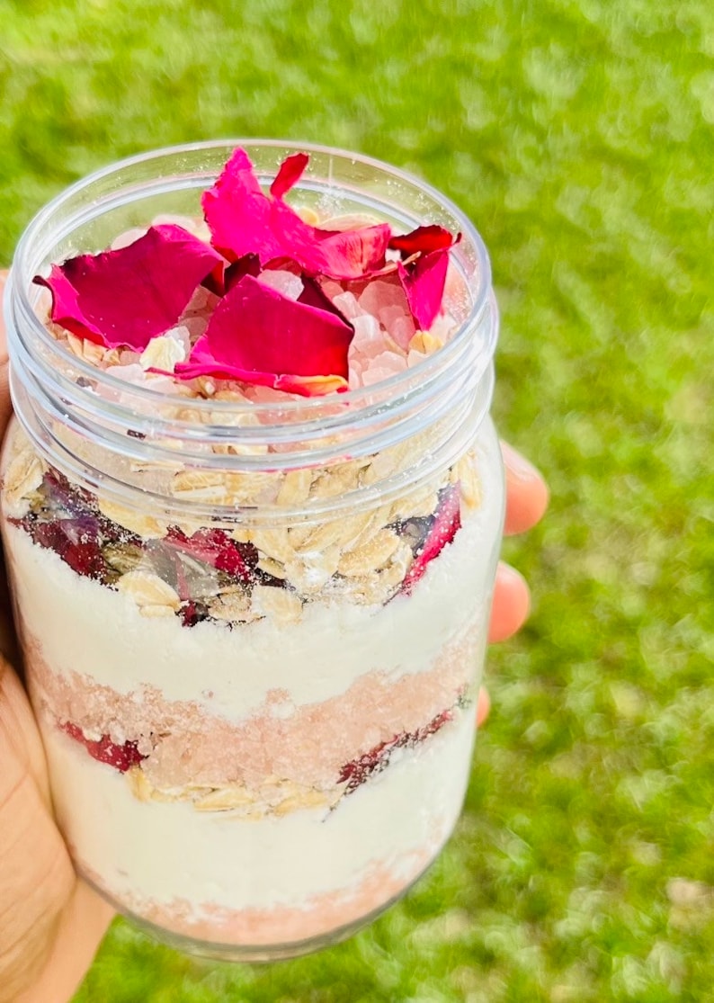 May include: A glass jar filled with layers of pink Himalayan salt, oatmeal, and rose petals. The jar is held in a hand against a green grassy background.