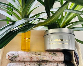 May include: A wooden tray holds a small yellow bottle, a silver tin, and three clear tubes filled with bath salts. The text "mom" and "Mother's Organic Magic" are printed in gold. The background features green leafy plants.