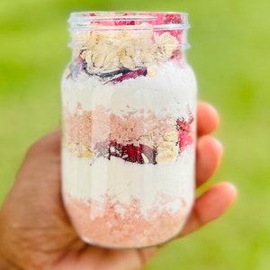 May include: A glass jar filled with layers of pink bath salts, white powder, dried rose petals, and oats.