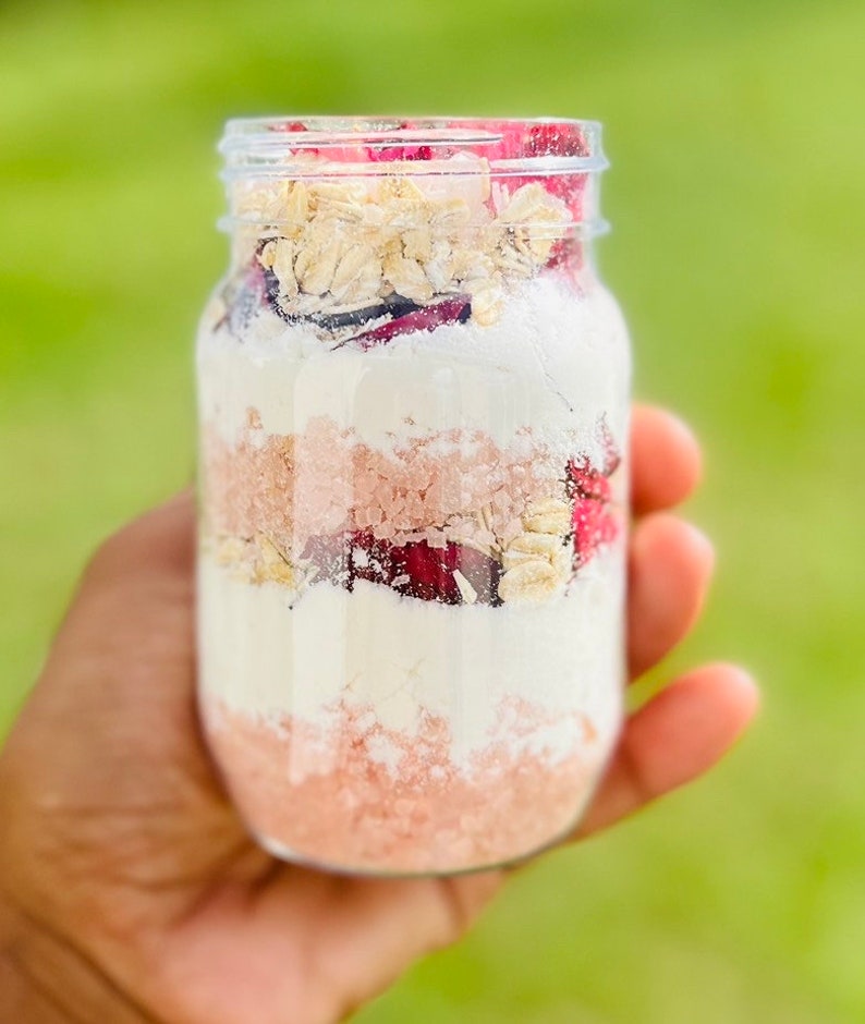 May include: A glass jar filled with layers of pink bath salts, white powder, dried rose petals, and oats.