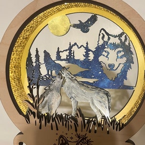 May include: A wooden wall decor with a circular cutout featuring a wolf howling at the moon, surrounded by trees and a flying eagle. The background is a starry night sky. The bottom of the decor has a wolf silhouette with floral accents.