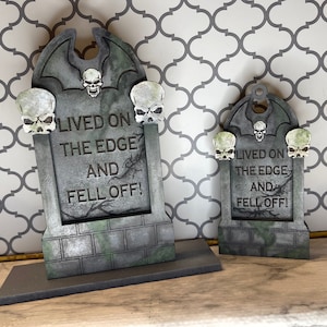 Op de afbeelding: Twee grijze en witte, grafsteen-vormige Halloween-decoraties met een vleermuisontwerp aan de bovenkant. De grotere grafsteen heeft een schedel aan de bovenkant en zegt "Lived on the edge and fell off!" De kleinere grafsteen heeft een schedel aan de bovenkant en zegt "Lived on the edge and fell off!"