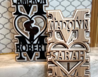 May include: Two wooden cutout letters "M" with names and hearts inside. The left "M" is black with white accents and the names "Cameron" and "Robert". The right "M" is natural wood with the names "Madolyn" and "Sarah" and "Mason" at the bottom.