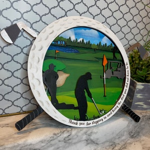Puede incluir: Una bola de golf de madera blanca con una escena de campo de golf verde en su interior. La bola de golf est&aacute; rodeada por dos palos de golf blanco y negro. El texto "Thank you for stepping in and being the dad you did not have to be" est&aacute; escrito en la parte inferior de la bola de golf.