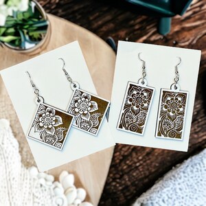 May include: Two pairs of wooden earrings with intricate floral designs. One pair features square-shaped earrings with a brown background and white floral designs. The other pair features rectangular earrings with a brown background and white floral designs.