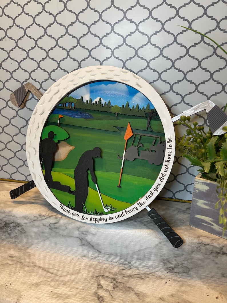 Puede incluir: Una bola de golf de madera blanca con una escena de campo de golf verde en su interior. La bola de golf est&aacute; rodeada por dos palos de golf negros con empu&ntilde;aduras blancas. El texto "Thank you for stepping in and being the dad you did not have to be." est&aacute; escrito alrededor del borde de la bola de golf.