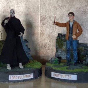 Może przedstawiać: Dwie figurki akcji, Lord Voldemort i Harry Potter, stoją na podstawach z kamiennym murem w tle. Voldemort jest ubrany w czarną szatę i trzyma różdżkę. Harry Potter jest ubrany w brązową kurtkę i dżinsy i trzyma różdżkę.